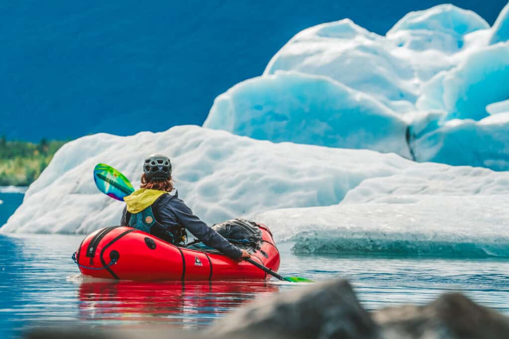 A man in a packraft paddling near a big iceberg in one of Alaska's glacier lagoons