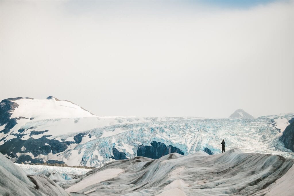 person stands on glacier in Alaska looking at massive hanging glacier in background